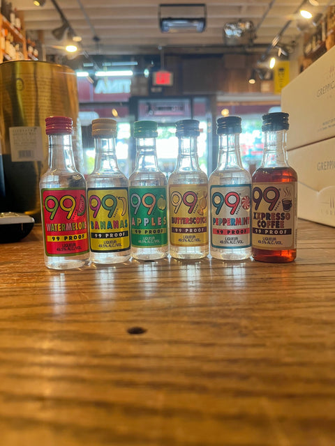 Small clear bottles with colorful labels that read 99, from left to right watermellon, bananas, apples, butterscotch, peppermint, and xpresso coffee, and 99 proof