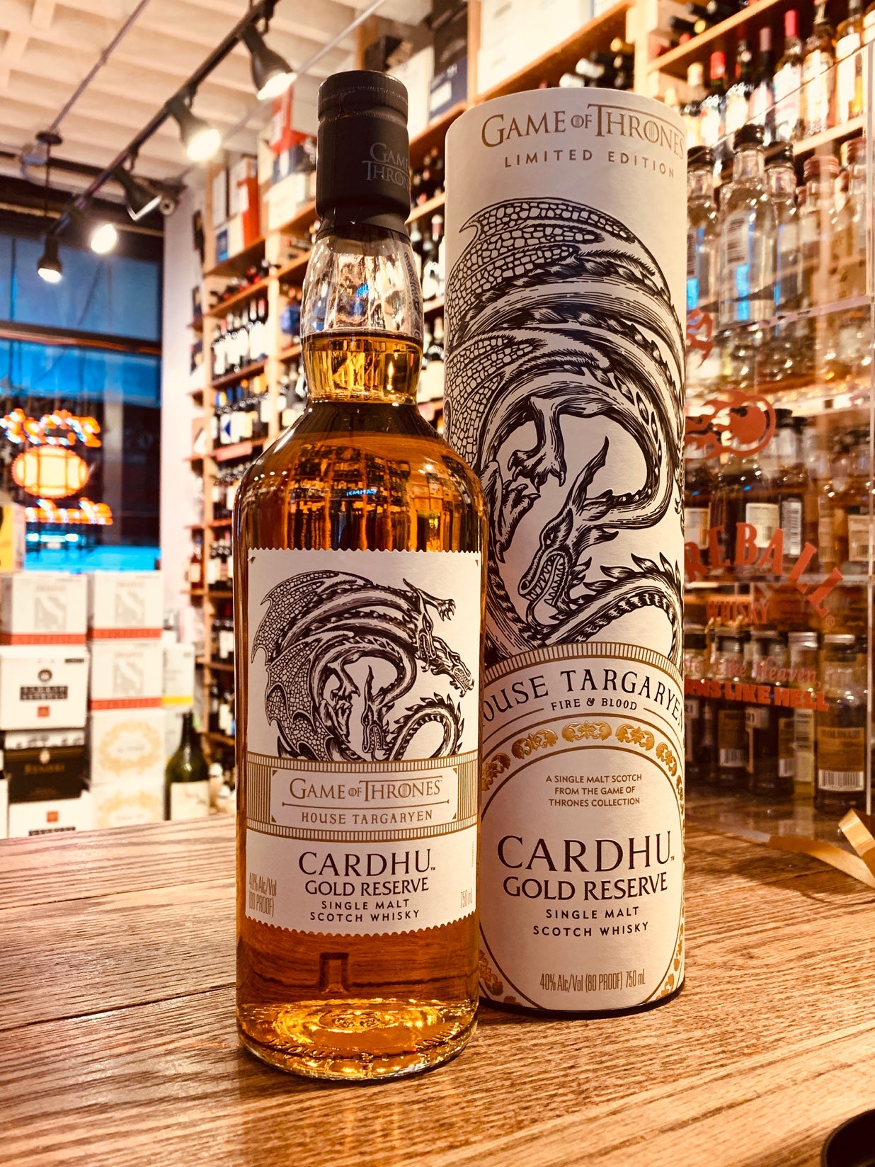 House Targaryan Cardhu Gold Reserve 750mL a tall clear glass bottle with a black top and white label with an image of a dragon on it next to a tall white tin cylinder with the image of a dragon on it