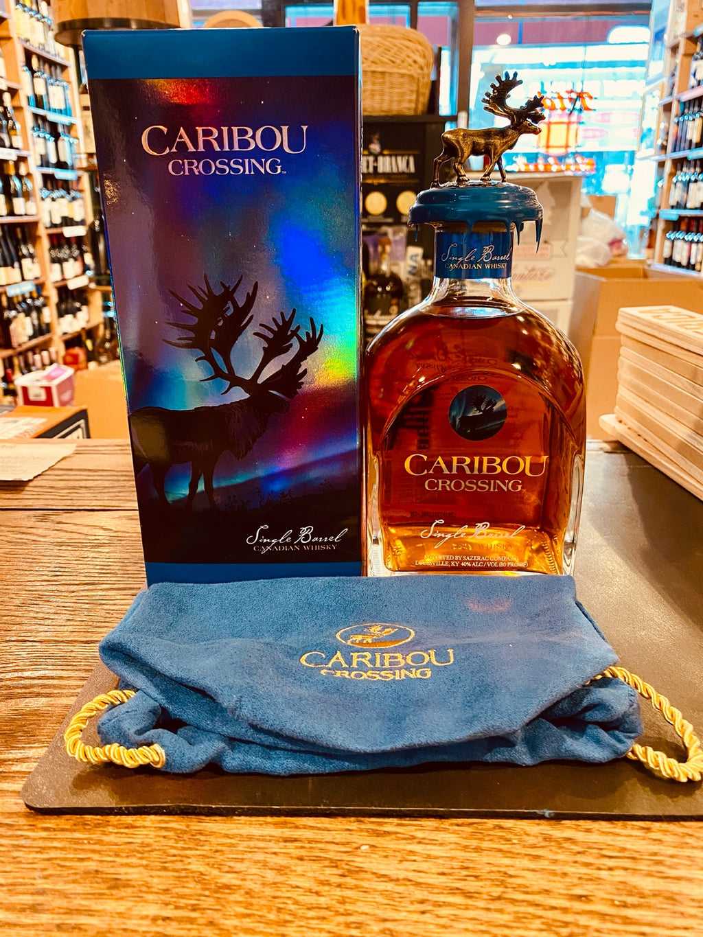 Caribou Crossing 750mL a blue reflective box with an image of a caribou on it next to a small square rounded squat clear bottle with gold lettering and a caribou emblem topper. a blue velvet sack lay in front of both