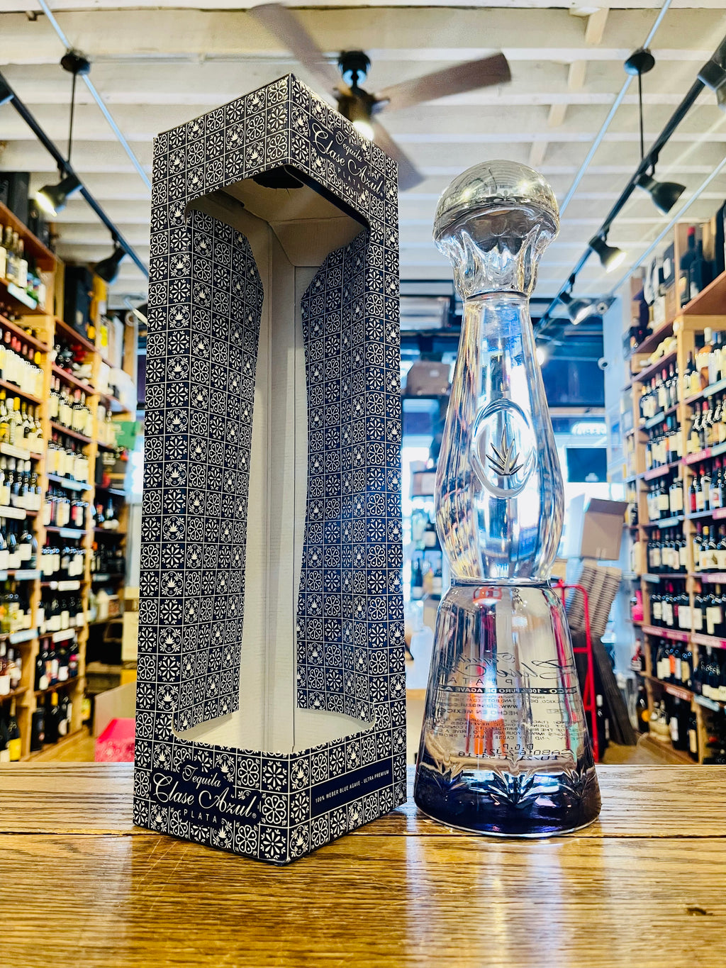 Clase Azul Plata 750 mL a tall elegantly designed clear bottle with a silver bell topper next to a tall blue and white box