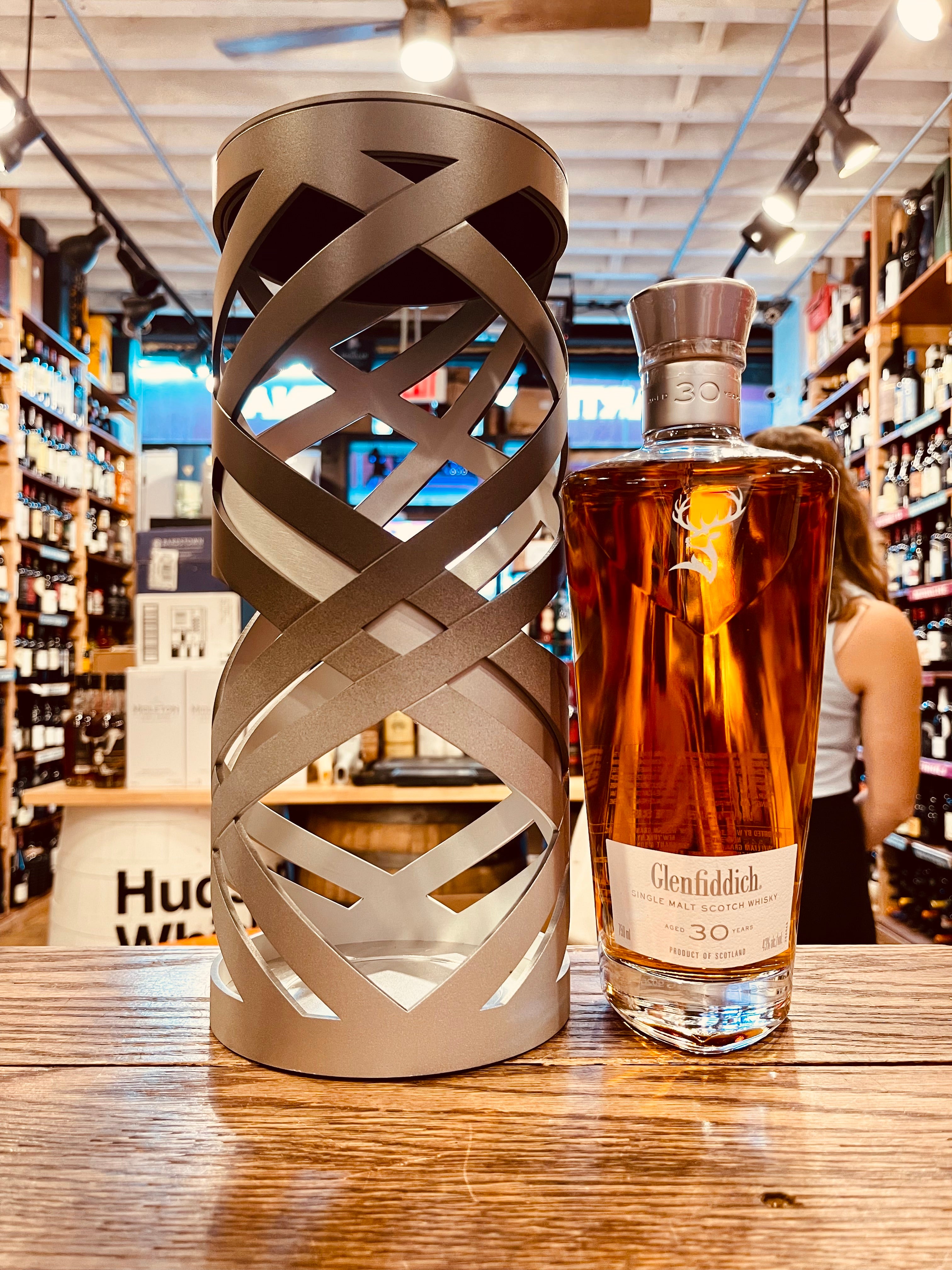 Glenfiddich 30Yr Suspended Time 750mL an open faced artistically designed metal silver  cylinder next to a clear angulated tall bottle with a white label and deer head on it