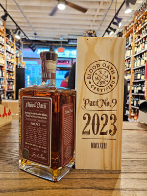 Blood Oath Bourbon Whiskey Pact No 9 750mL a squared clear bottle with red labeling, next to a wooden box that has burnt in lettering 