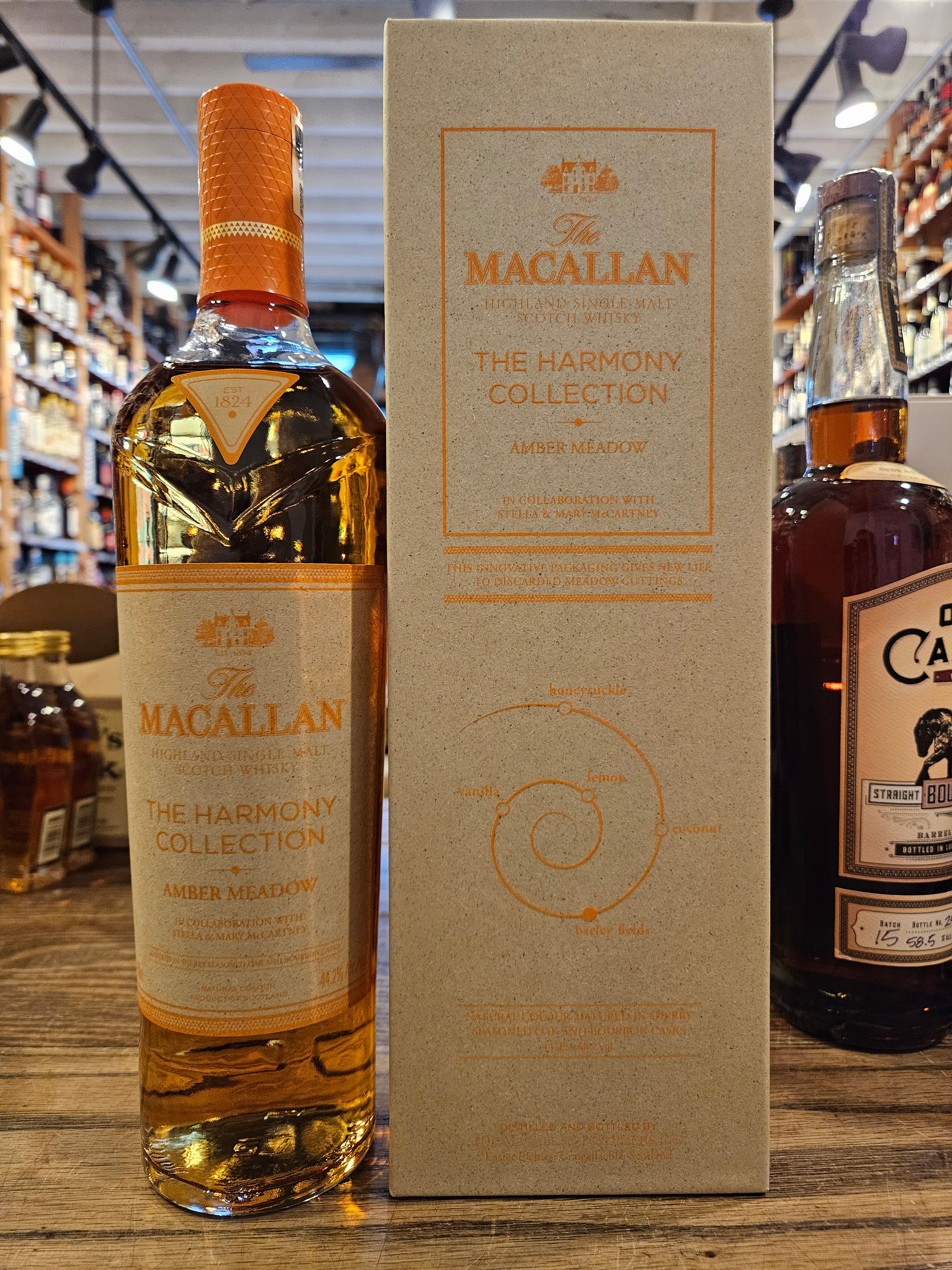 Macallan Harmony Collection Amber Meadow 750mL a tall slender clear glass bottle with a gray and orange label with an orange top next to a gray and orange box