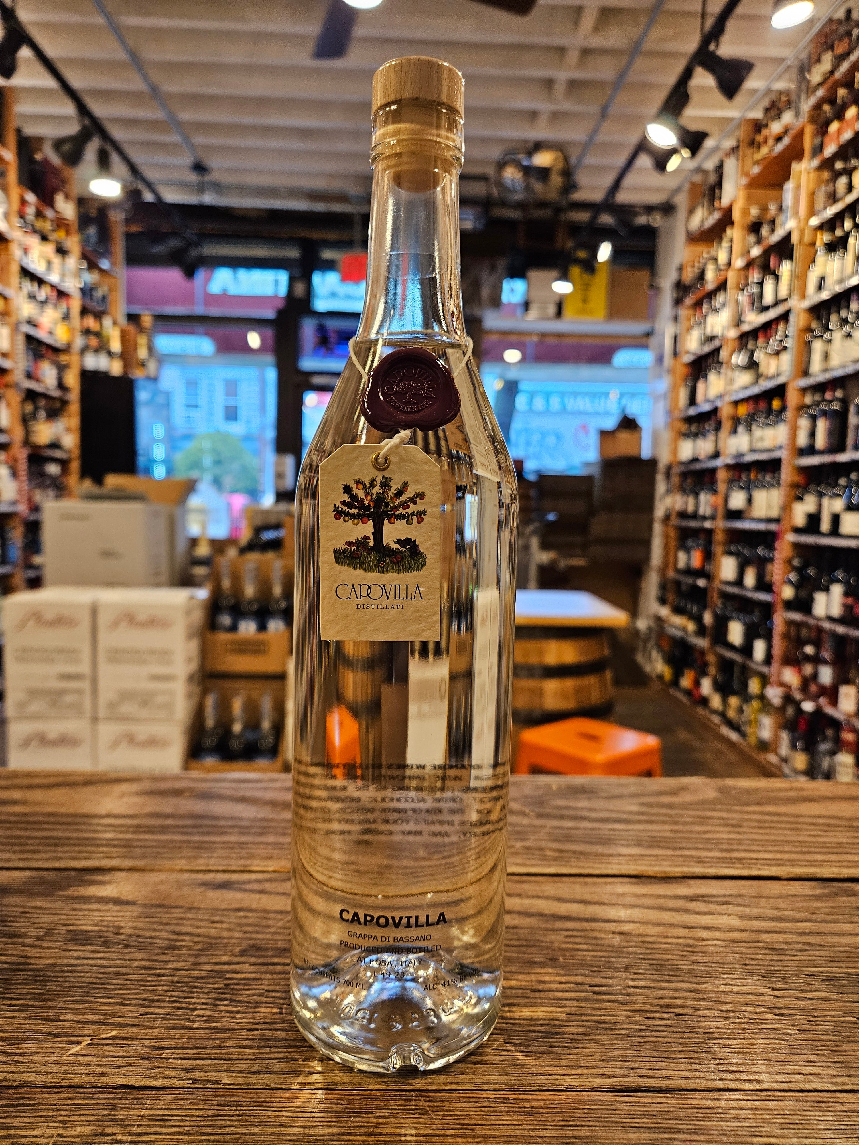 Capovilla Distillati Grappa di Bassano 700mL a slender clear glass bottle with a tapered neck and a beige tag hanging around the bottle neck with a maroon wax stamp on it and a wood top