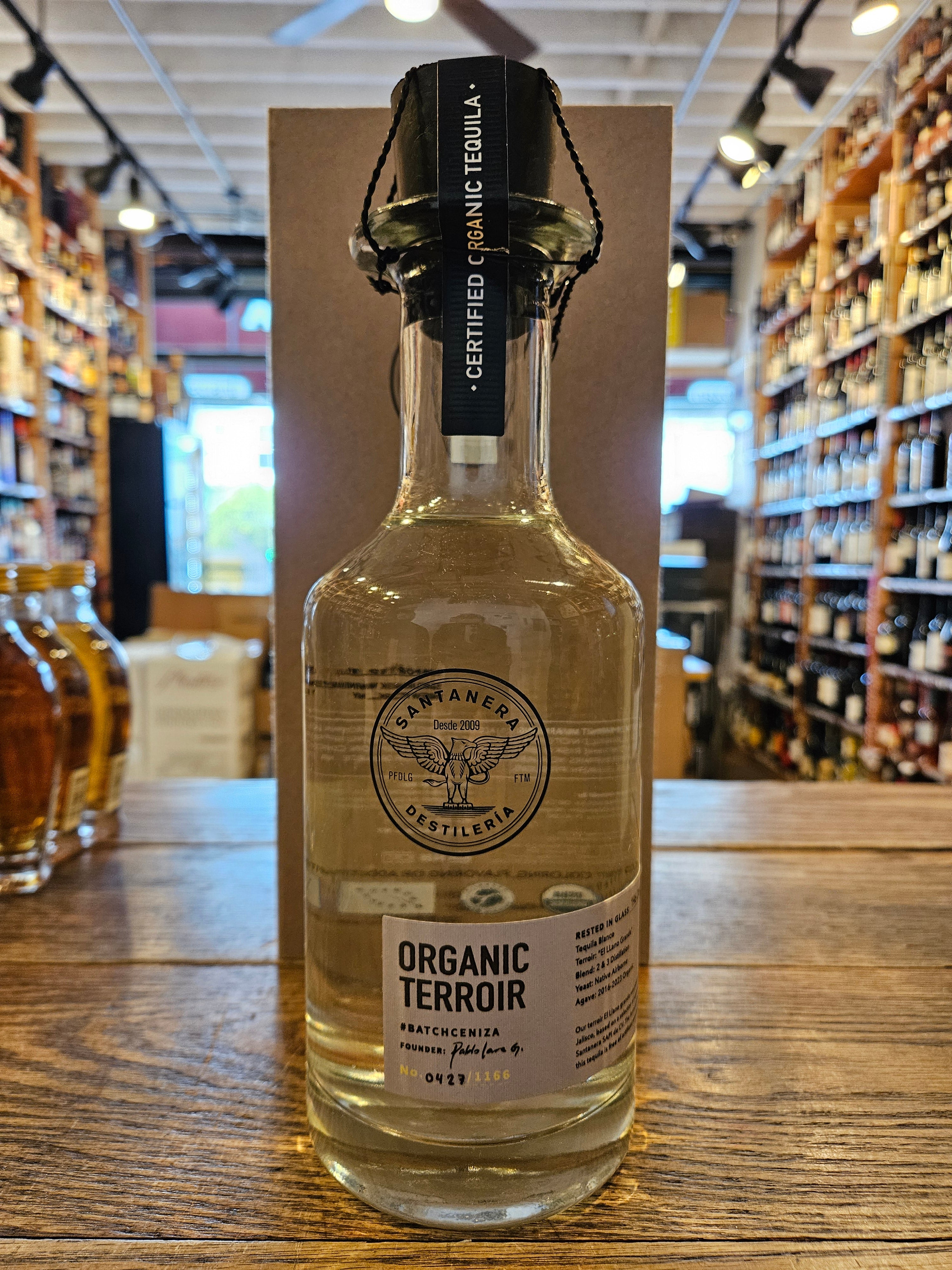 Santanera Organic Tequila Blanco Batch-Ceniza 750mL a clear round shouldered glass bottle with a thick neck and white label and black wooden top in front of a rectangular brown box