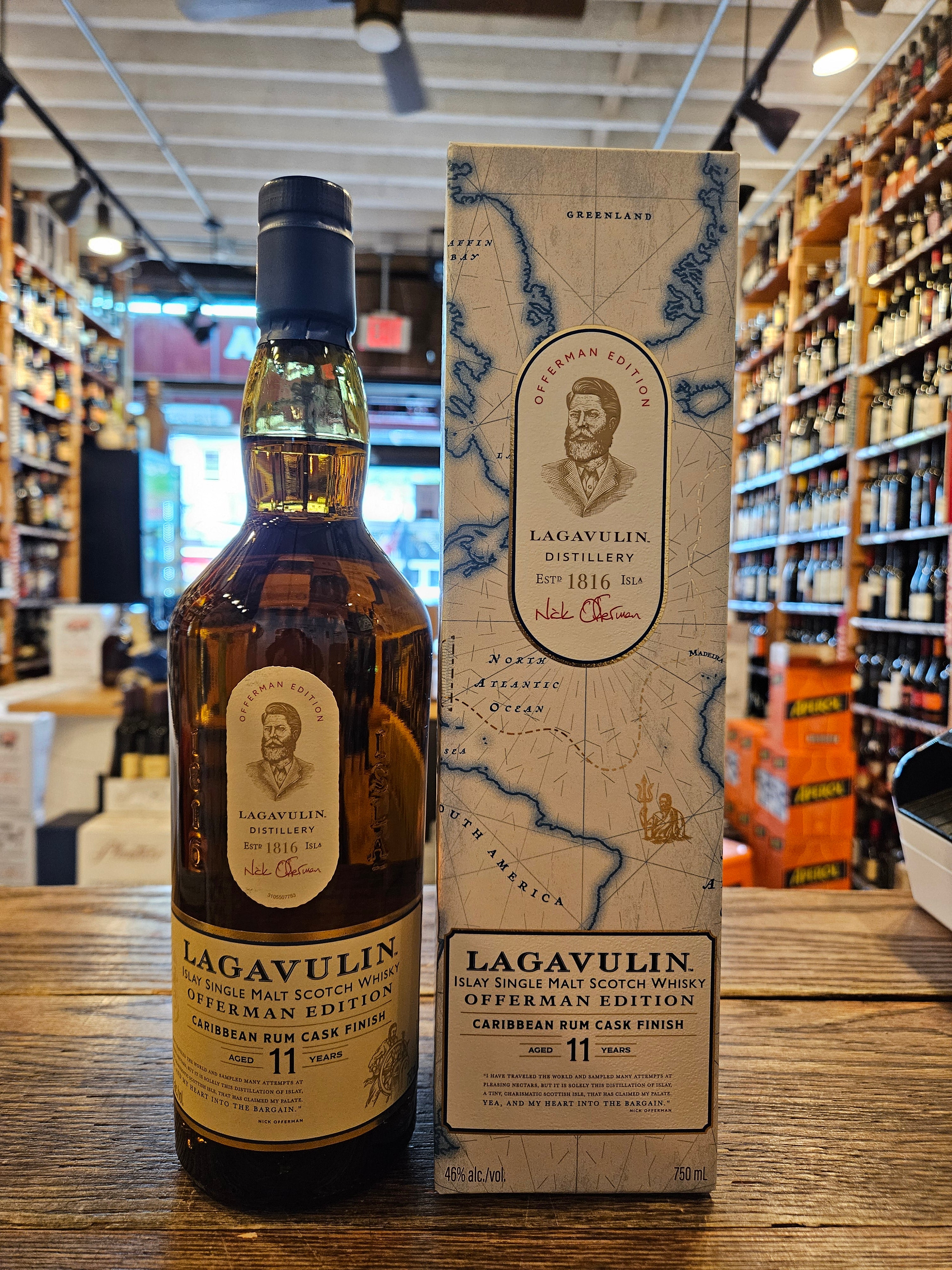 Lagavulin 11 Year Offerman Edition Caribbean Rum Cask Finish Single Malt Scotch Whisky a clear glass bottle with a beige label and a blue top next to a tall rectangular white box with an atlas on it.