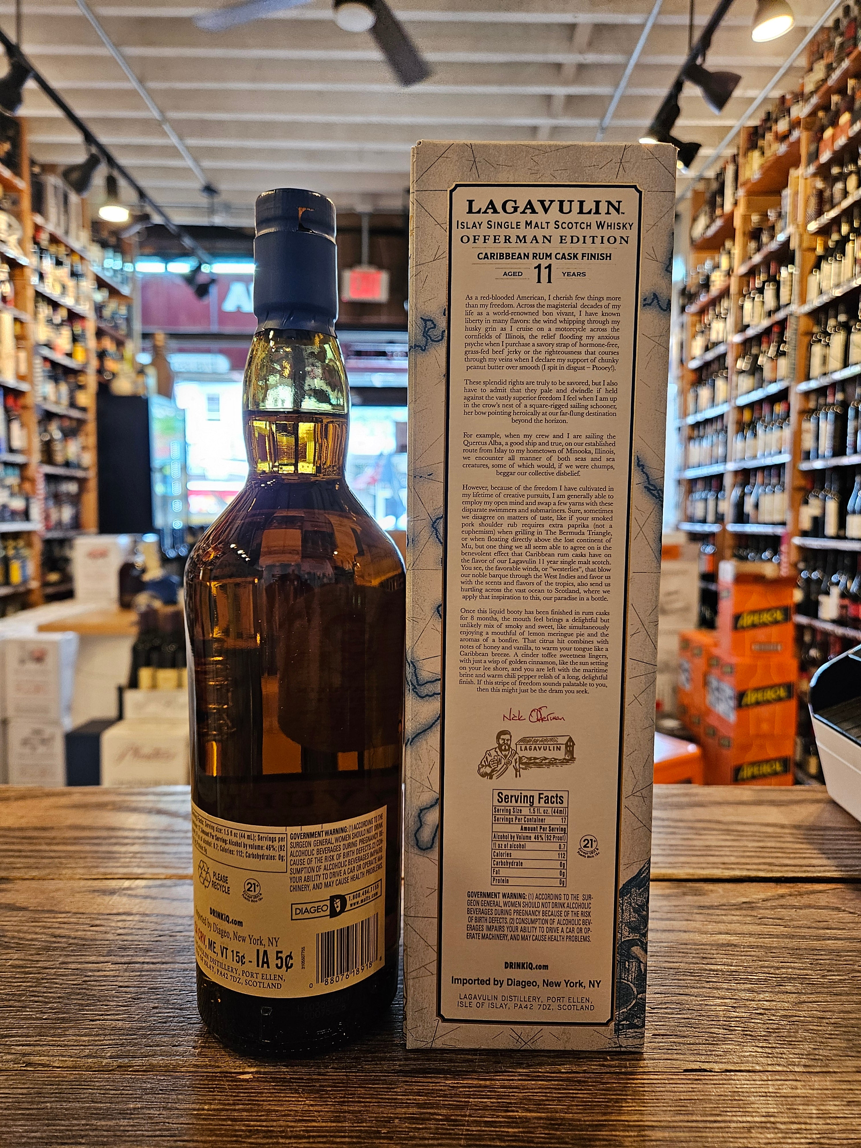 Lagavulin 11 Year Offerman Edition Caribbean Rum Cask Finish Single Malt Scotch Whisky 750mL the backside of a clear glass bottle with a beige label and a blue top next to a tall rectangular white box with an atlas on it.