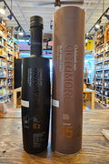 Octomore 15.2 700mL a dark black frosted glass bottle with a slender neck next to a tall brown colored cylinder.