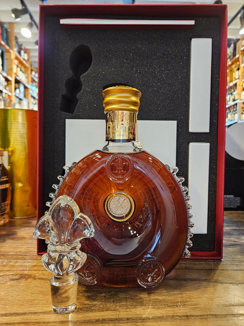 Louis XIII 750mL a round crystal clear glass bottle with a golden top and a crystal glass topper, in front of a red box with black padding