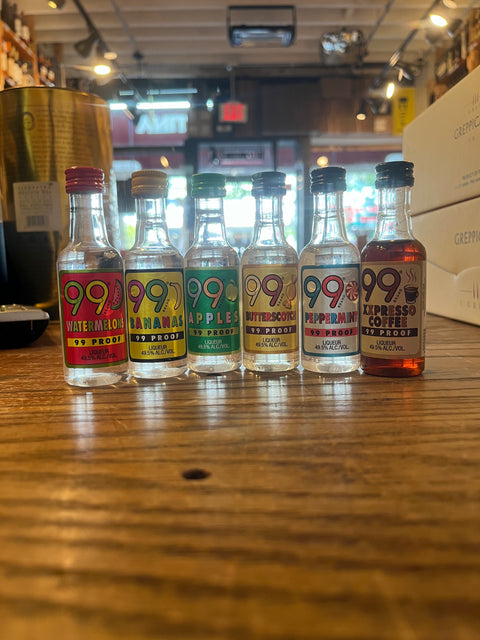 Small clear bottles with colorful labels that read 99, from left to right watermellon, bananas, apples, butterscotch, peppermint, and xpresso coffee, and 99 proof