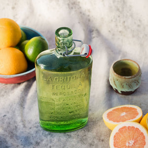 La Gritona Reposado 750mL a clear green flat glass bottle with a pop top next to a bowl with oranges and limes 