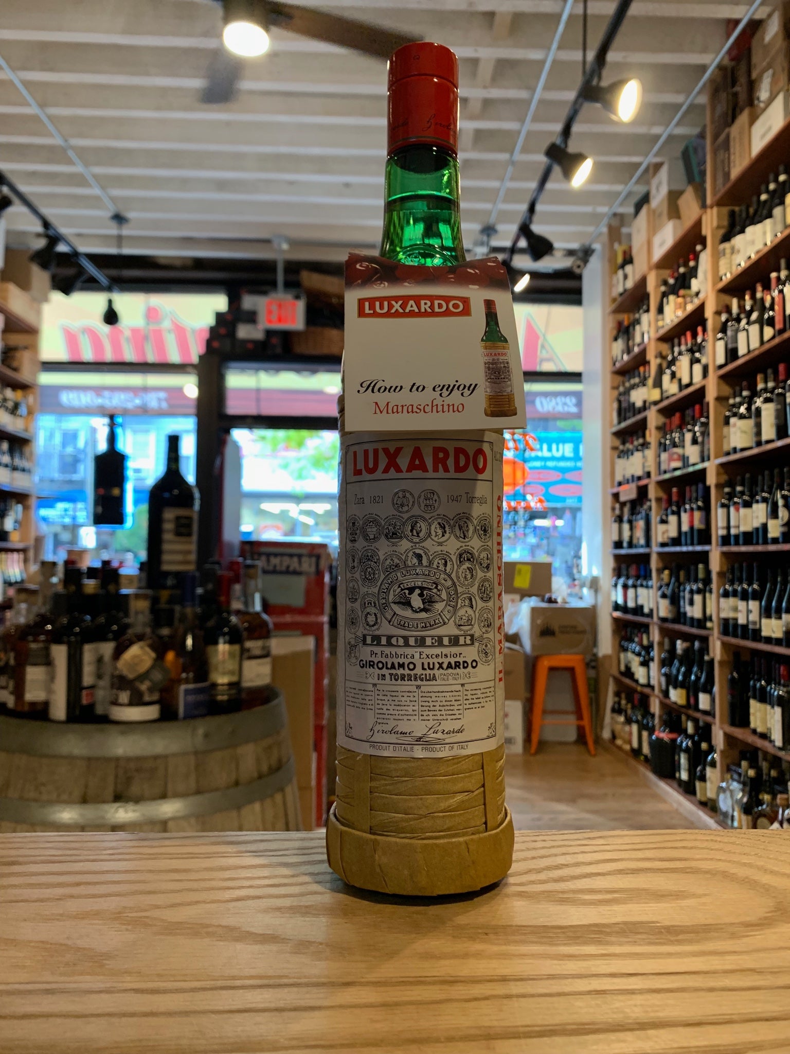 Luxardo Maraschino Liquer 64 750mL a tall slender clear green glass bottle with a twine basket around the bottle and white label and red top