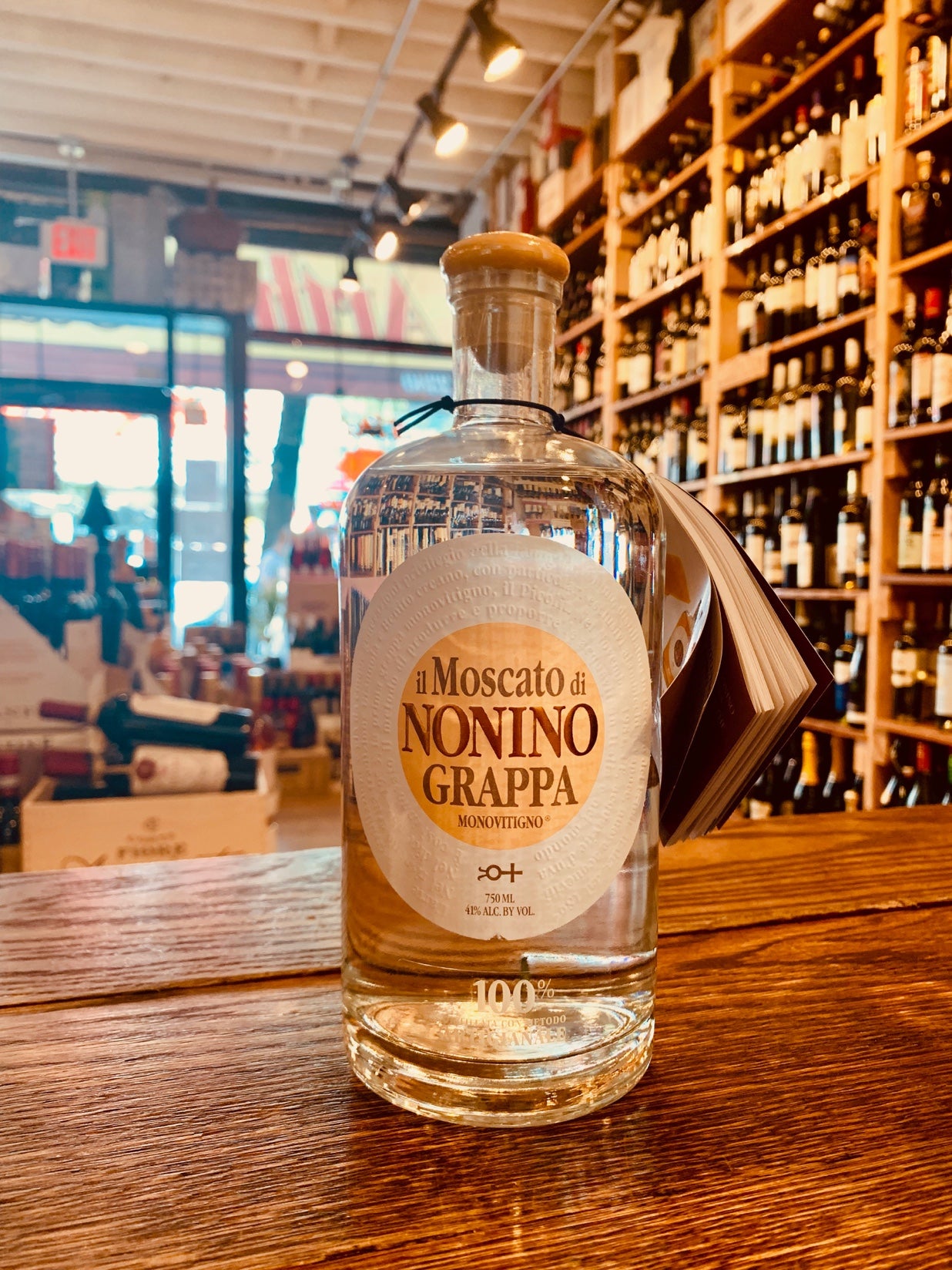 Grappa Nonino Moscato 750mL a short squat round shouldered clear bottle with a stubby neck and white label and wooden top