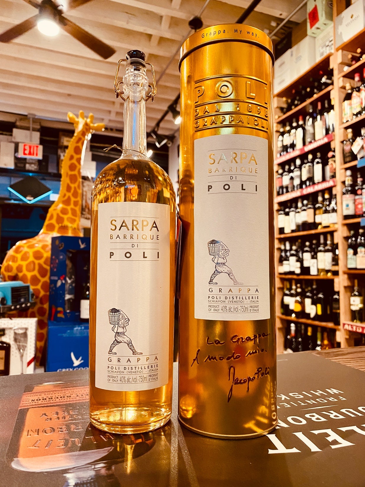 Jacopo Poli Barrique Grappa 750mL a tall slender clear glass bottle with a slender neck and a large white label next to a tall golden color tin cylinder with a white label 
