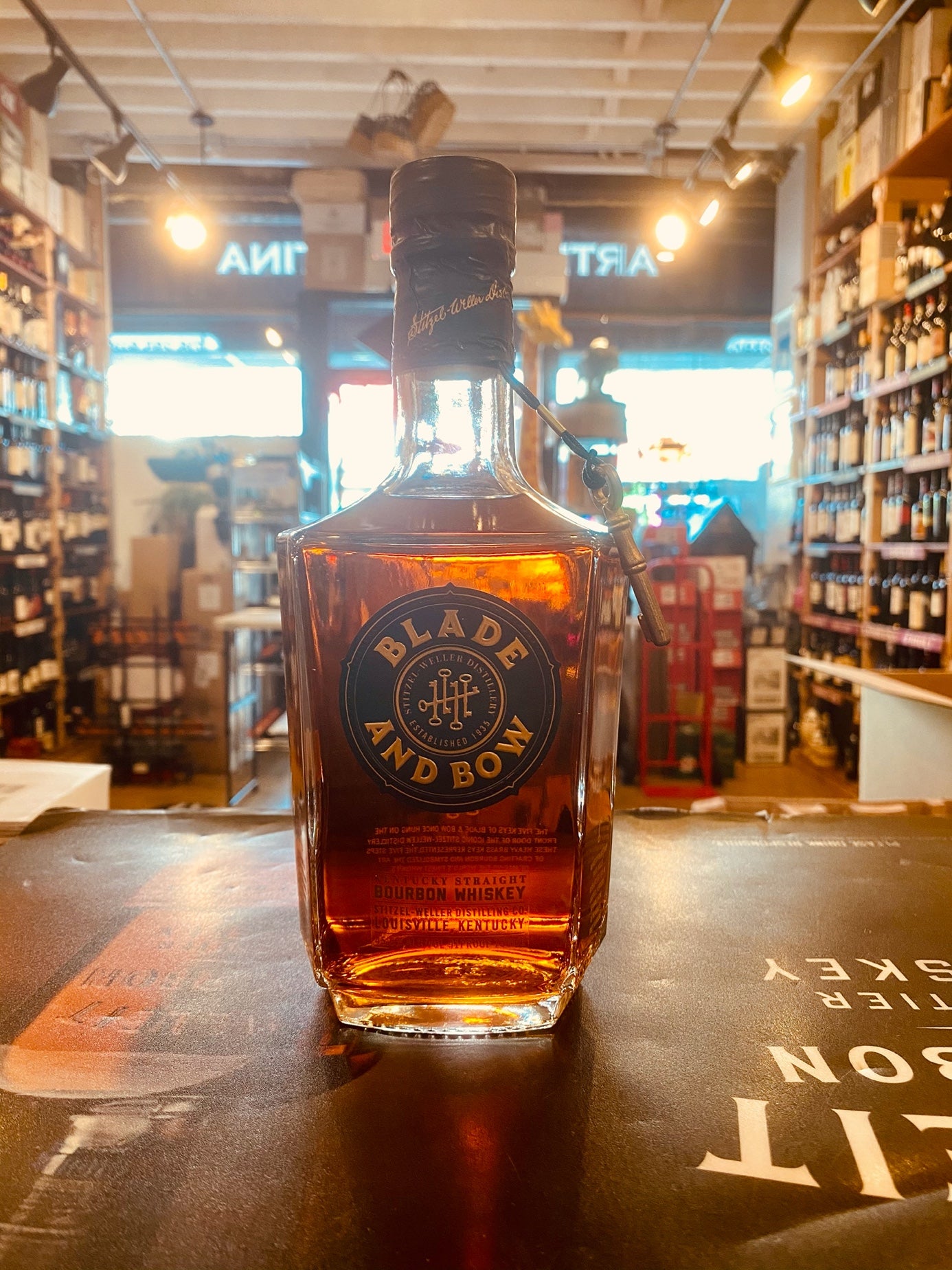 Blade & Bow Bourbon 750mL small squared squat bottle with a dark blue and gold label and a key amulet hanging off the neck