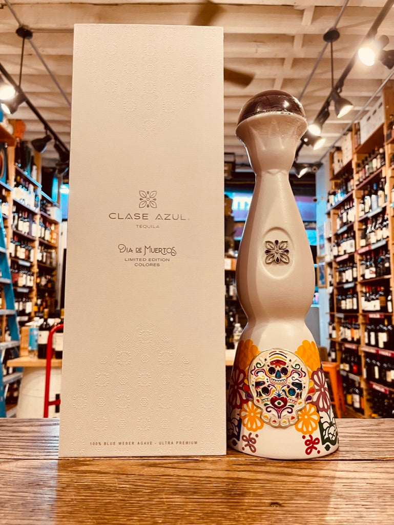 Clase Azul Dia De Los Muertos 1L a tall elegantly designed white bottle with colorful skull design and a silver bell topper next to a tall white box with silver lettering