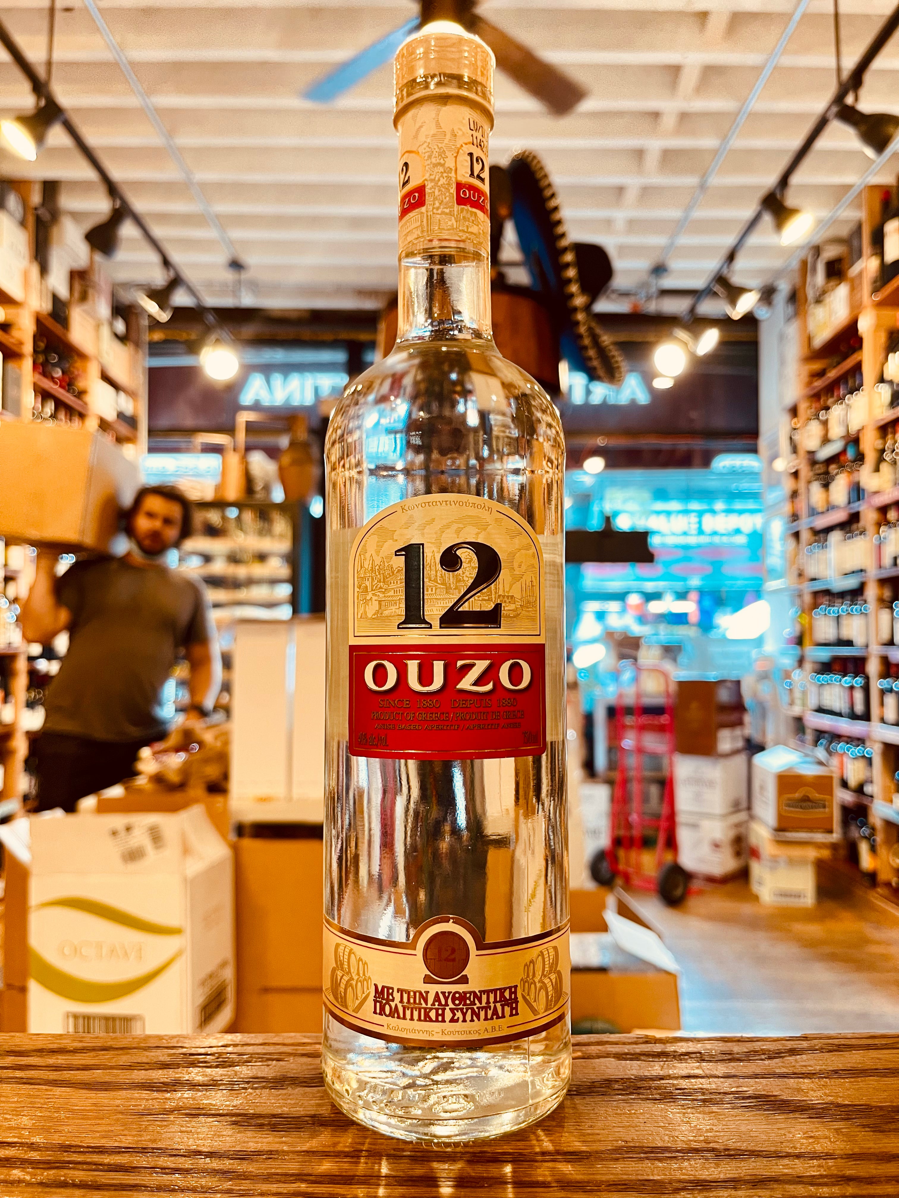 Ouzo 12 750mL a tall clear glass bottle with a white and red label and wooden top