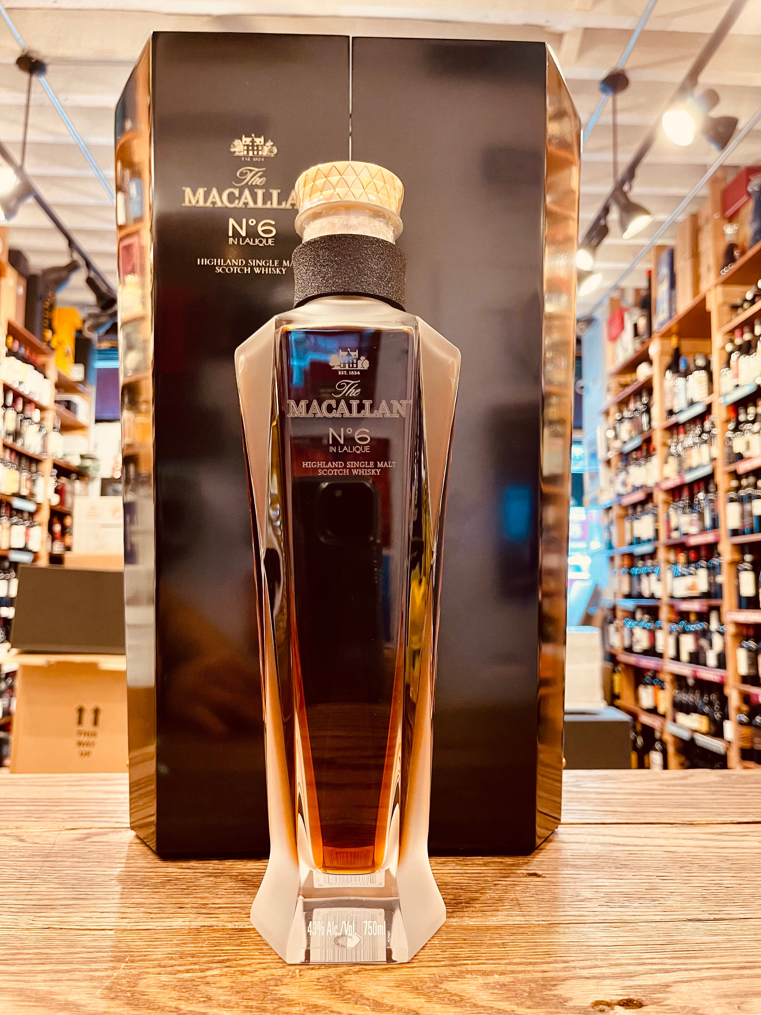 The Macallan Decanter Series V6 'No. 6 in Lalique' 750mL a tall hexagon shaped black box behind a tall elegant hexagon shaped clear glass bottle with gold lettering and a glass top