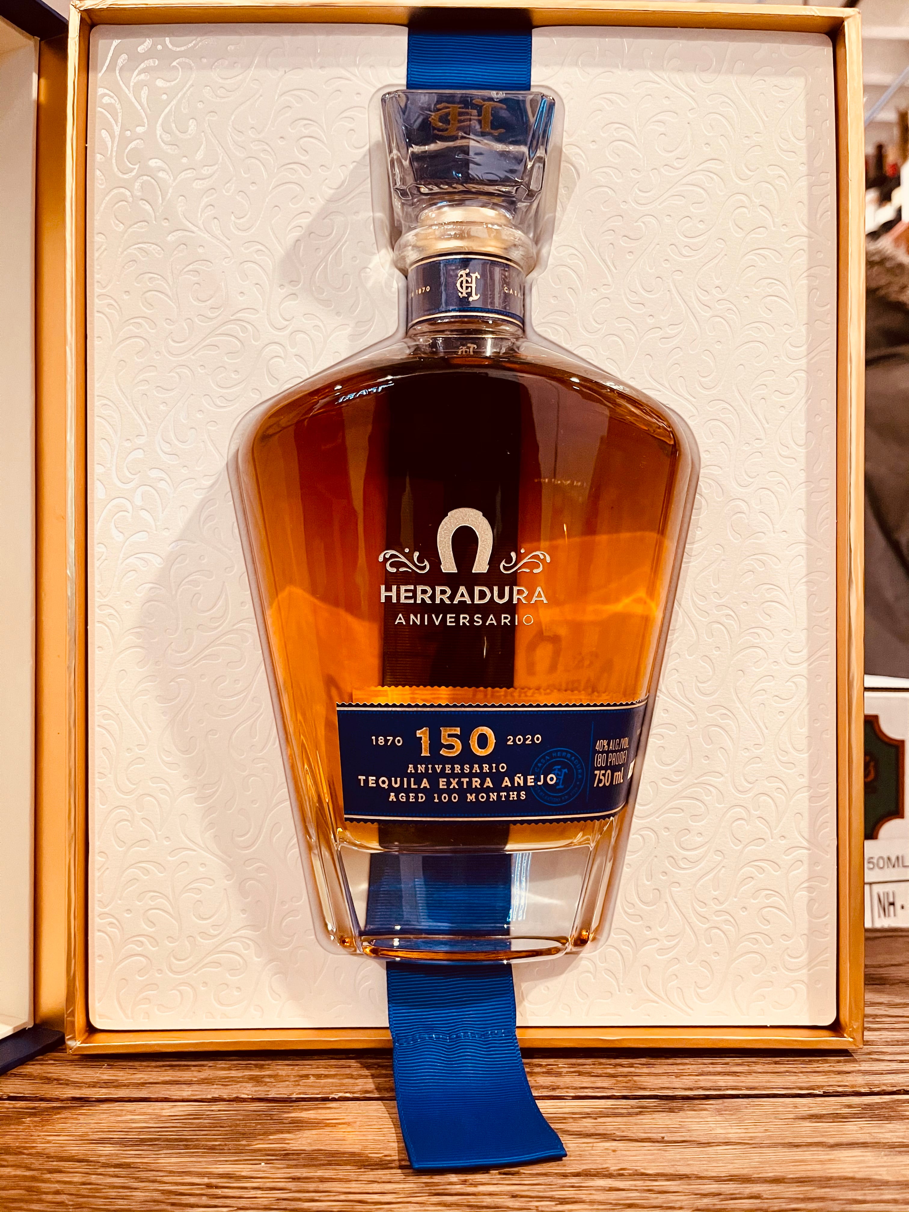 Herradura Aniversario 750mL an open box with white lining holding a shapely wide shouldered clear glass bottle with a blue label and silver lettering and a glass top