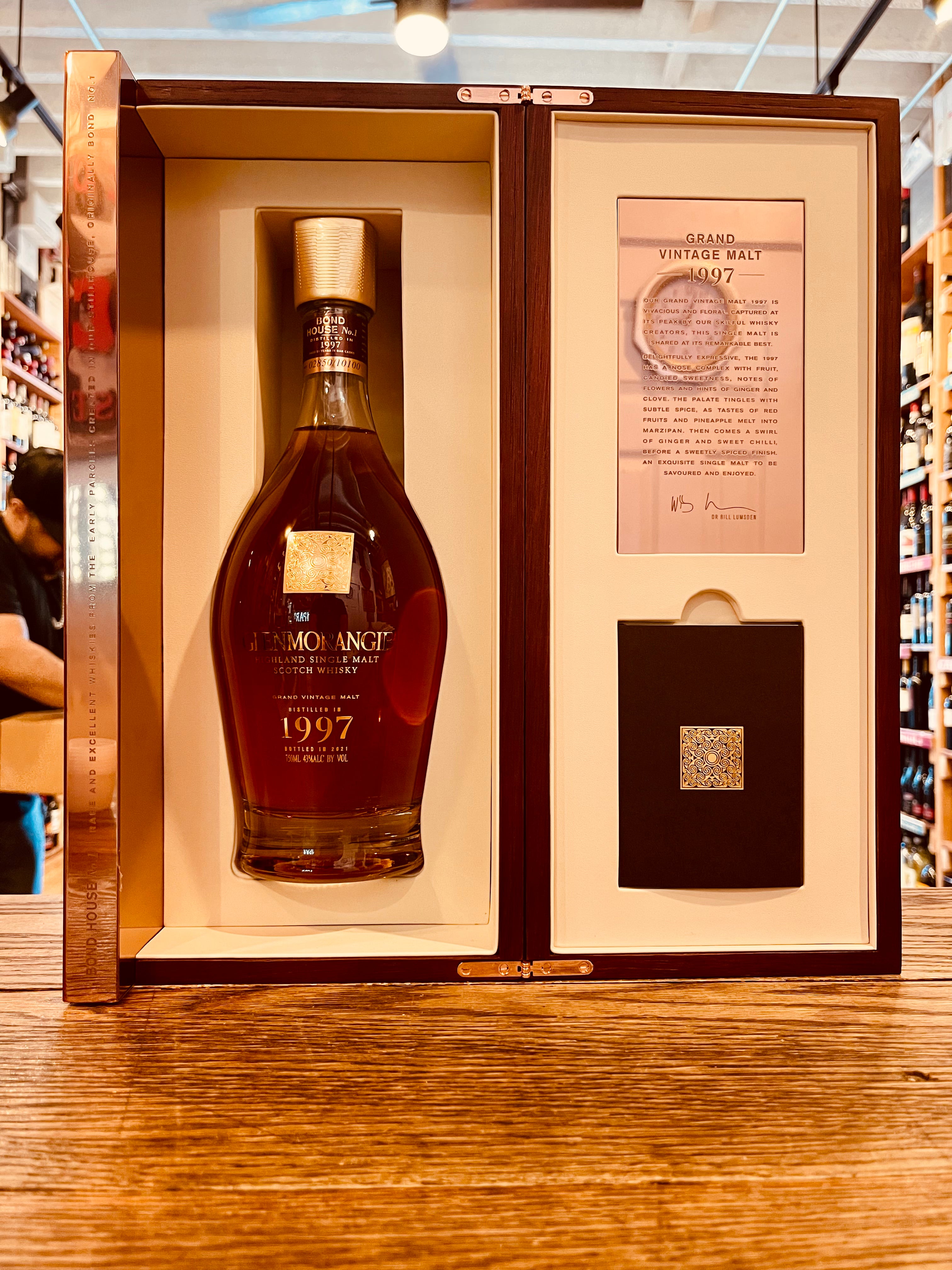 Glenmorangie 1997 Bond House No.1 750mL an open wooden gift box with white lining and a robust rounded clear bottle  with golden lettering and gold top and pamphlet inside