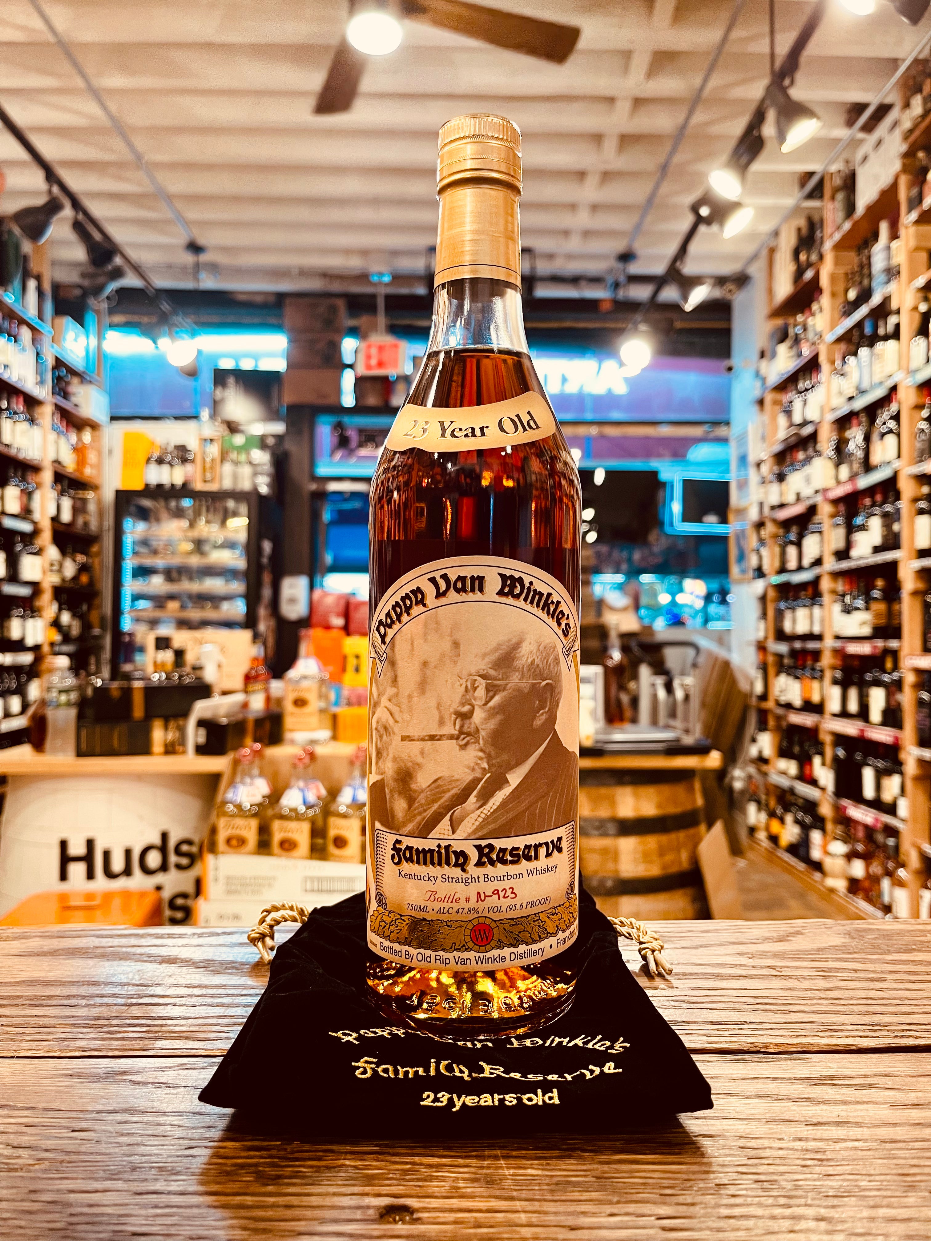 Pappy Van Winkle 23Yr 750mL a tall clear glass bottle with a tapered neck with a beige label and an image of an old man smoking a cigar on it and a golden top, the bottle is sitting on a black velvet sack that has golden font
