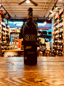 Quilt Napa Cabernet Reserve 750mL 2017 a dark tall glass wine bottle with a black and gold label and a black top