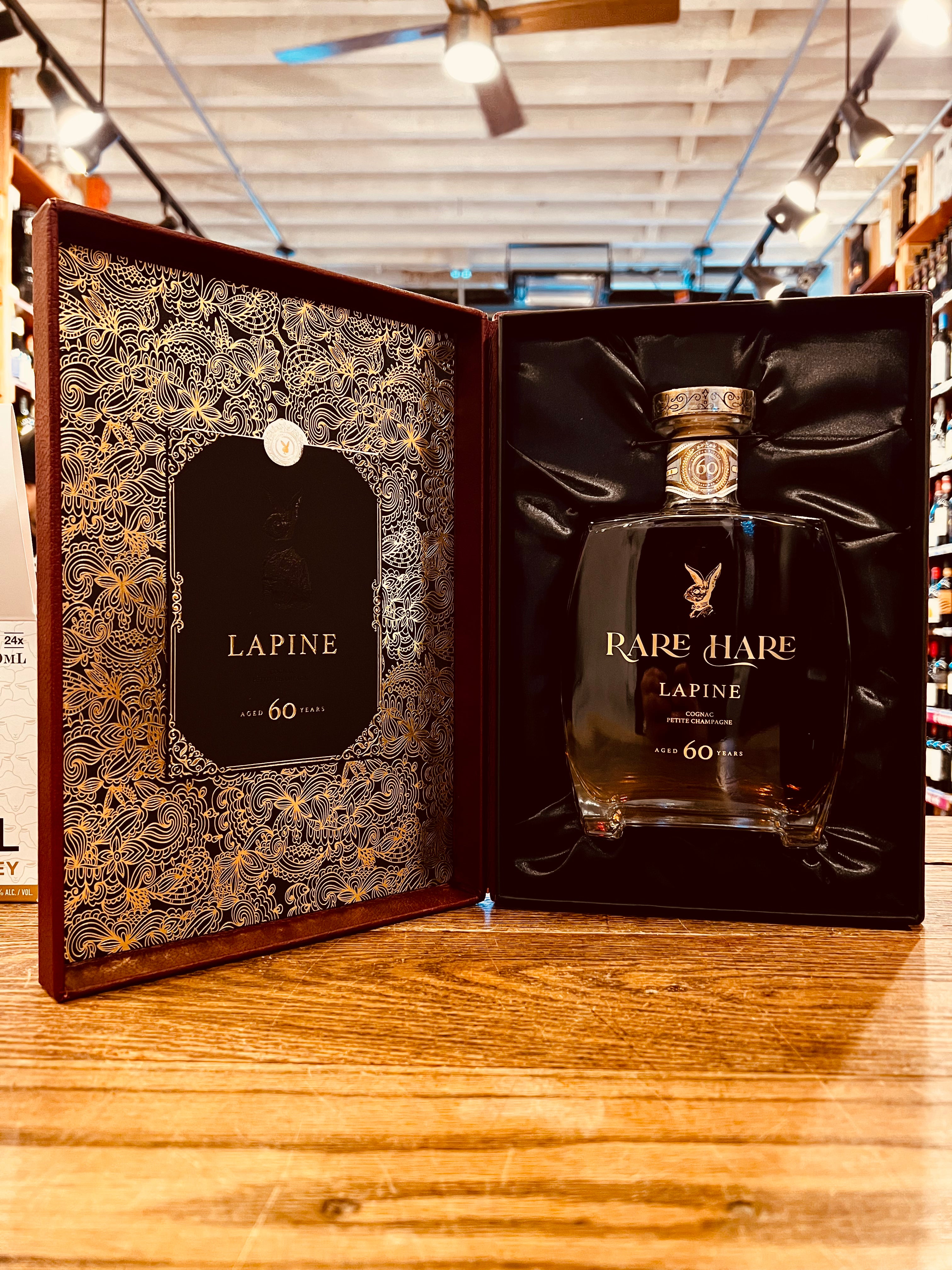 Rare Hare Cognac Lapine Aged 60 Years 750mL an open box one side lined in a golden intricate design the other sided being lines in black velvet holding a short squared clear glass bottle with golden lettering and a golden top 