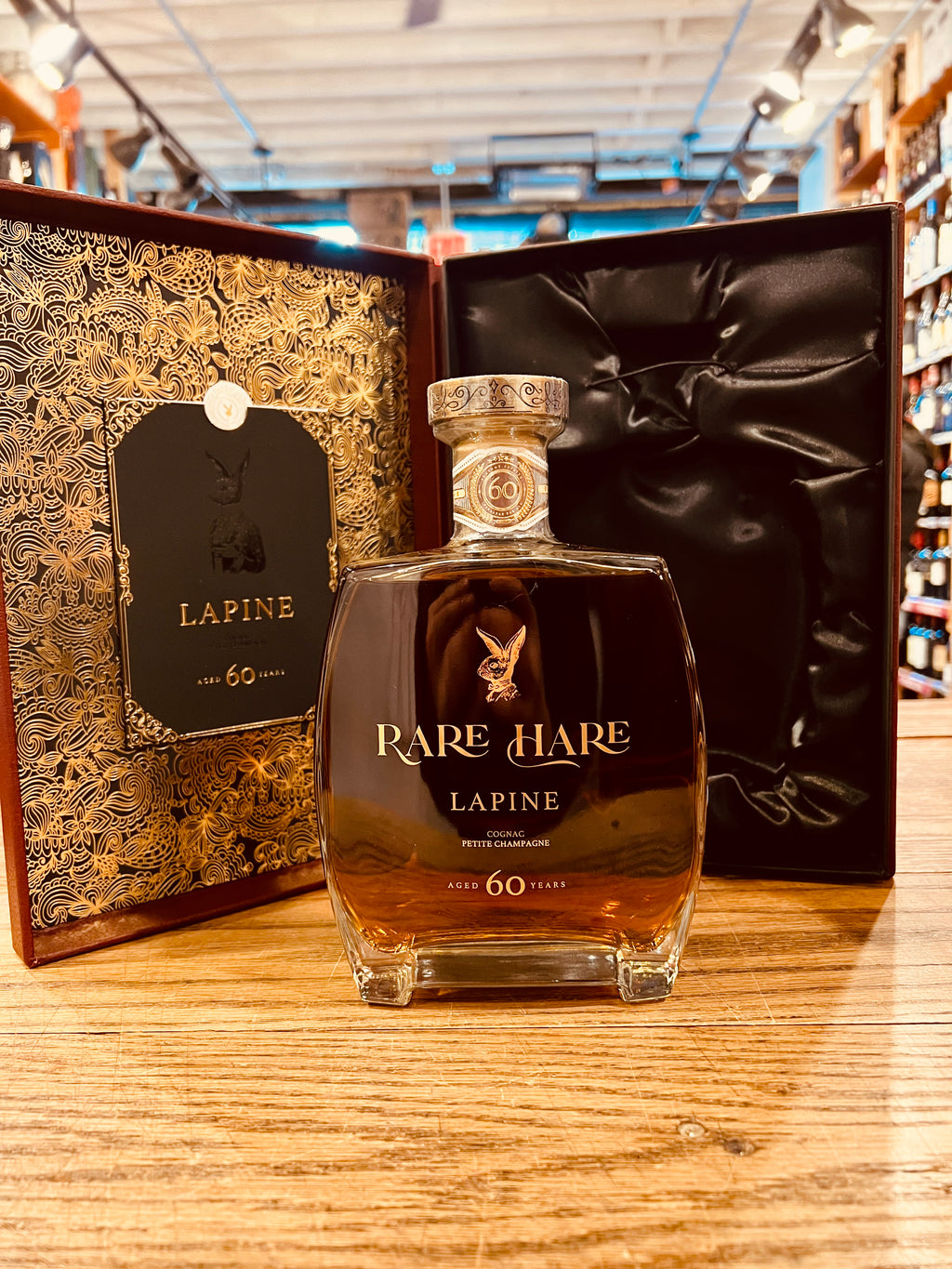 Rare Hare Cognac Lapine Aged 60 Years 750mL an open box with one side being lined in black velvet while to other being lined in an intricate golden design with a squared short glass bottle with gold lettering and a golden top in front of the open box
